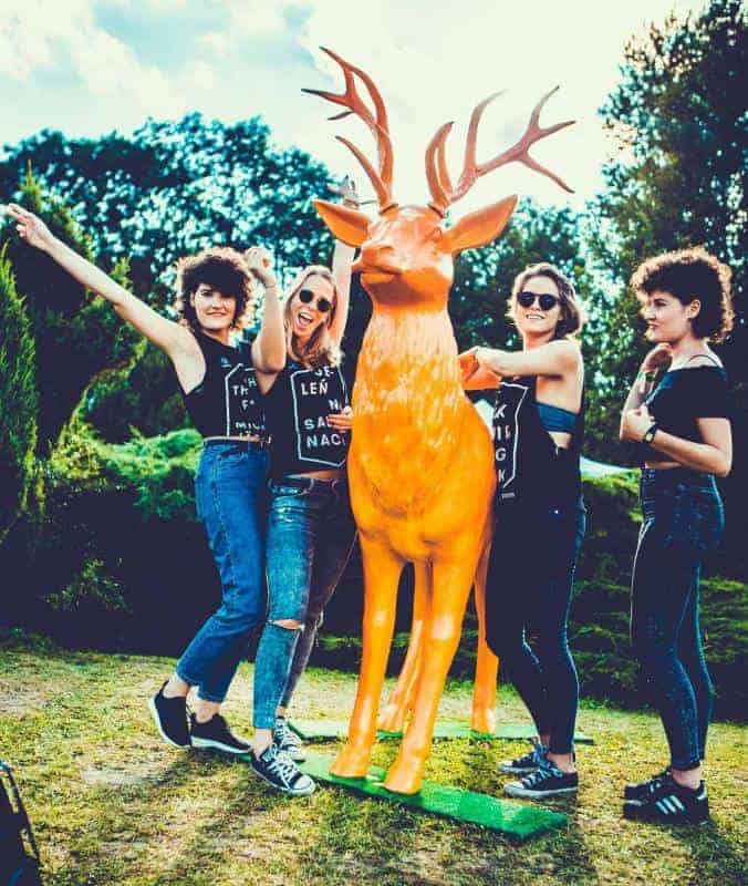 Hirsch Jägermeister 2 Cztery kobiety pozują przy pomarańczowej figurze jelenia na tle zieleni.