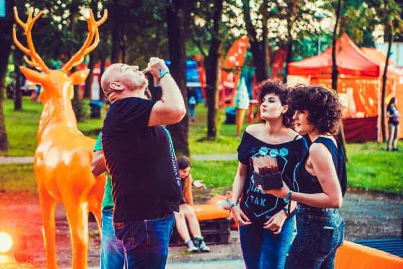 Hirsch Jägermeister 4 Ludzie na festiwalu na świeżym powietrzu, z dużą pomarańczową figurą jelenia w tle, w otoczeniu drzew i stoisk.