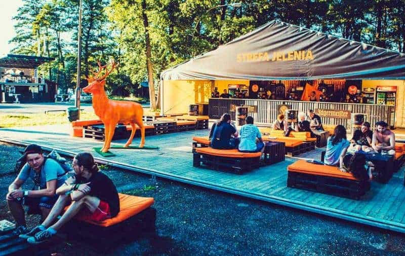 Hirsch Jägermeister 1 Ludzie siedzący na pomarańczowych poduszkach na drewnianej platformie przed namiotem z napisem