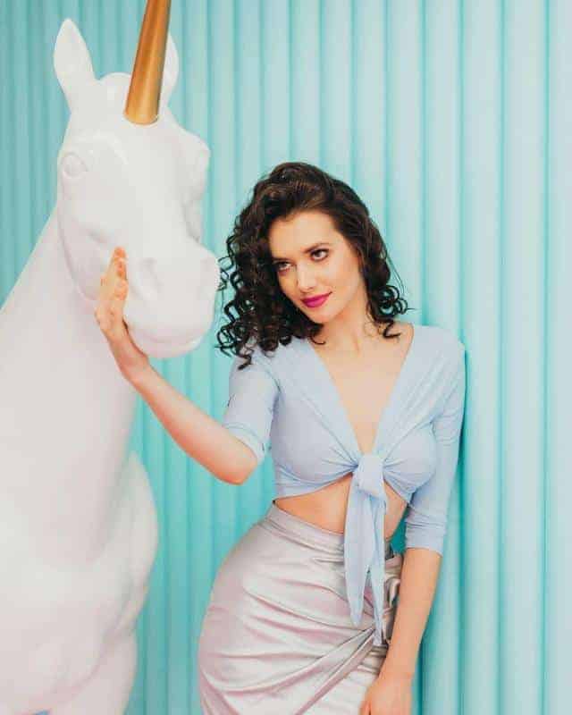 Die Ausstattung eines Fotostudios 2 Kobieta w błękitnej bluzce i srebrnej spódnicy, stojąca obok białej rzeźby jednorożca na tle turkusowej ściany.