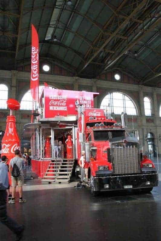 100. Jubiläum der Coca-Cola-Glasflasche 3 Czerwony ciężarówka Coca-Cola z naczepą przekształconą w stoisko sprzedażowe z produktami Coca-Cola, stojąca w dużej hali.