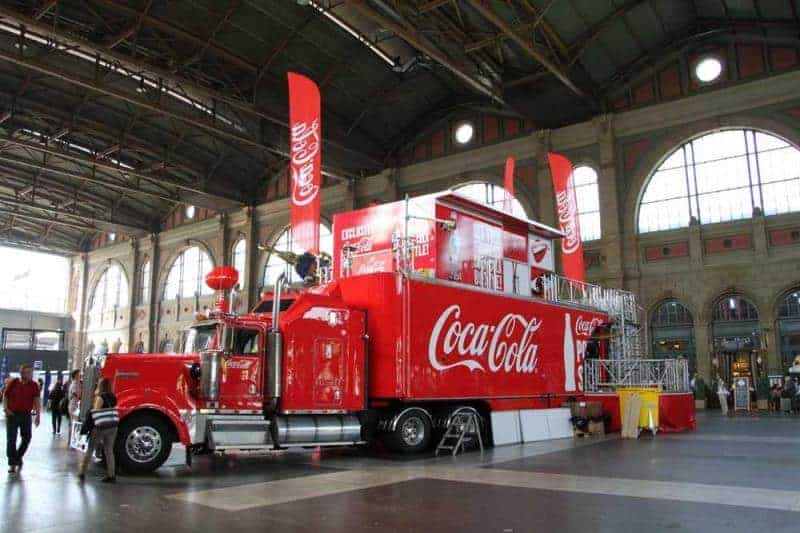 100. Jubiläum der Coca-Cola-Glasflasche 4 Czerwony ciężarówka Coca-Cola w hali dworca z dużymi flagami Coca-Cola i dużymi oknami w tle.