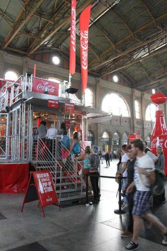100. Jubiläum der Coca-Cola-Glasflasche 2 Stoisko Coca-Coli z ludźmi w hali dworca kolejowego.