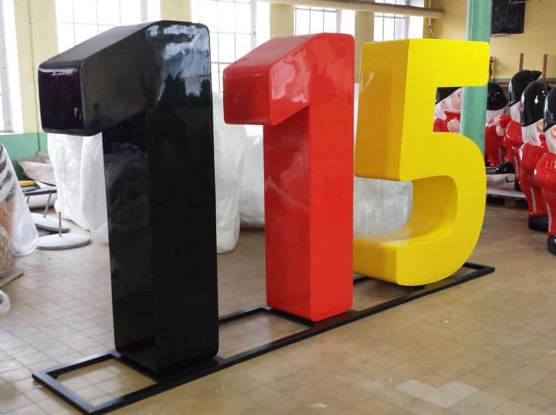 Czarno-czerwono-żółte rzeźby liczb 1, 1 i 5 na stalowej podstawie w pomieszczeniu.