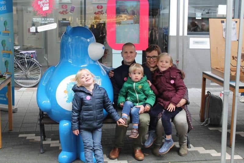 Bär auf einer Bank 2 Rodzina z trójką dzieci pozuje do zdjęcia obok dużej niebieskiej maskotki w przestrzeni publicznej.