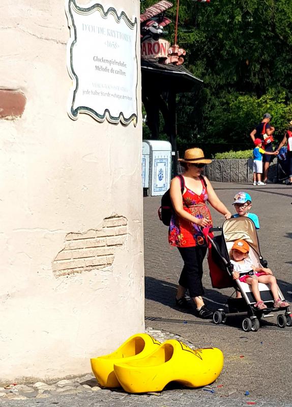 Große Holzschuhe 4 Kobieta z wózkiem dziecięcym przechodzi obok dużych żółtych drewniaków przy ścianie z francuskim napisem.