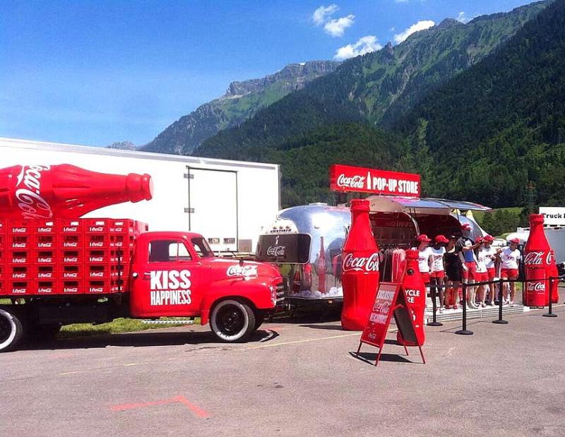 Werbetruck von Coca Cola 3 Czerwony samochód Coca-Coli z dużą butelką na przyczepie i sklepem pop-up w tle, stojący na parkingu w otoczeniu gór.