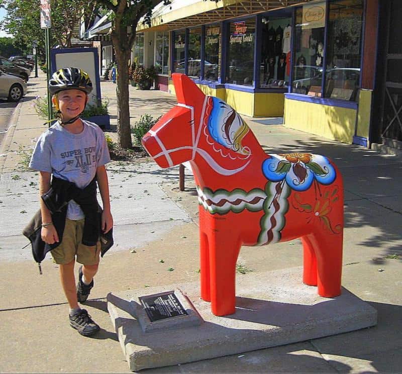 Künstlerisch bemalte Pferde aus Dalarna 2 Chłopiec w kasku na rower stojący obok kolorowej rzeźby konia na chodniku wzdłuż ulicy z witrynami sklepów.