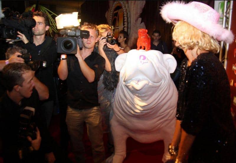 Kobieta w różowym kapeluszu pozuje na czerwonym dywanie obok dużej, białej rzeźby psa, otoczona przez fotoreporterów.