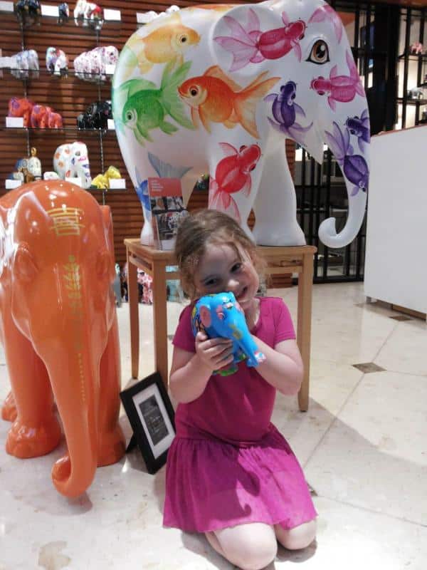 Elefantenparade 3 Dziewczynka w różowej sukience, trzymająca kolorową figurkę słonia, siedząca przy większych, pomalowanych słoniach dekoracyjnych.