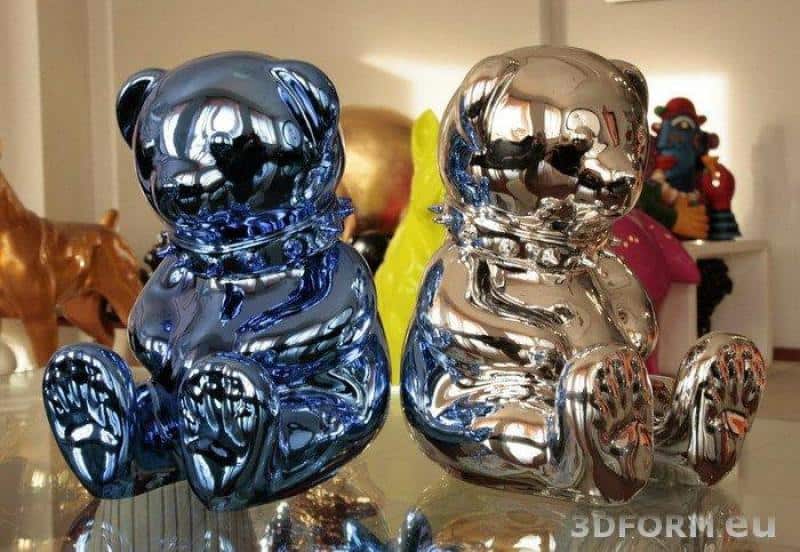 Metallisierte, verchromte Figuren 3 Dwie błyszczące figurki misiów, jedna w kolorze niebieskim, druga srebrna, stojące na szklanej powierzchni.