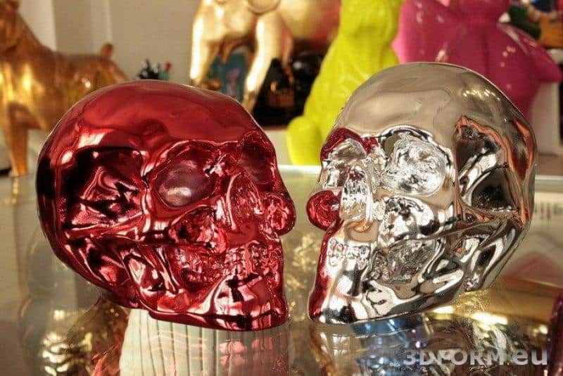 Metallisierte, verchromte Figuren 4 Dwie metaliczne czaszki, jedna czerwona, druga srebrna, ustawione na szklanym stole.