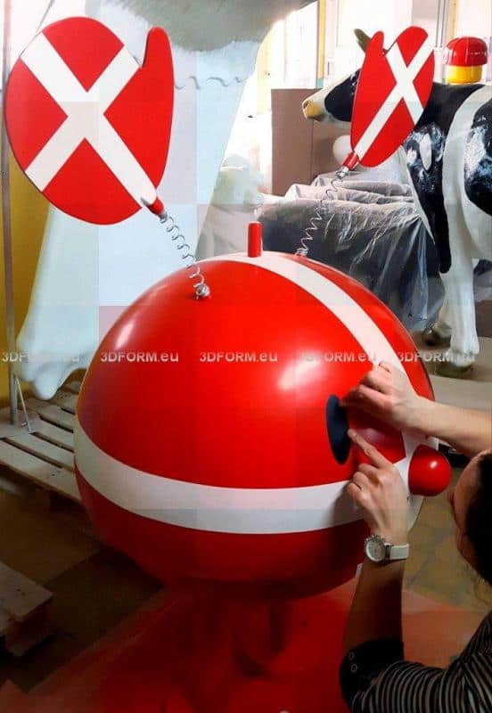 Czerwona, okrągła figura z uszami na sprężynach i białymi paskami, z rękami przymocowującymi oko. W tle plastikowa krowa.