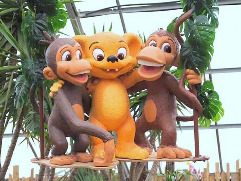 Trzy postacie kreskówkowe: dwa małpy i jeden stwór o pomarańczowej sierści, stoją razem na huśtawce w otoczeniu roślin.