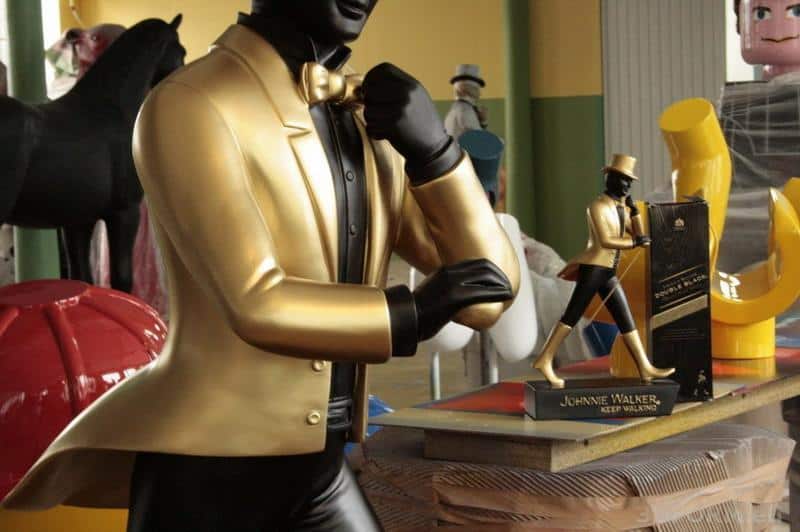 Große Johnnie Walker Figuren 1 Złota figurka Johni Walker w złotym garniturze, obok mniejsza statuetka i pudełko.