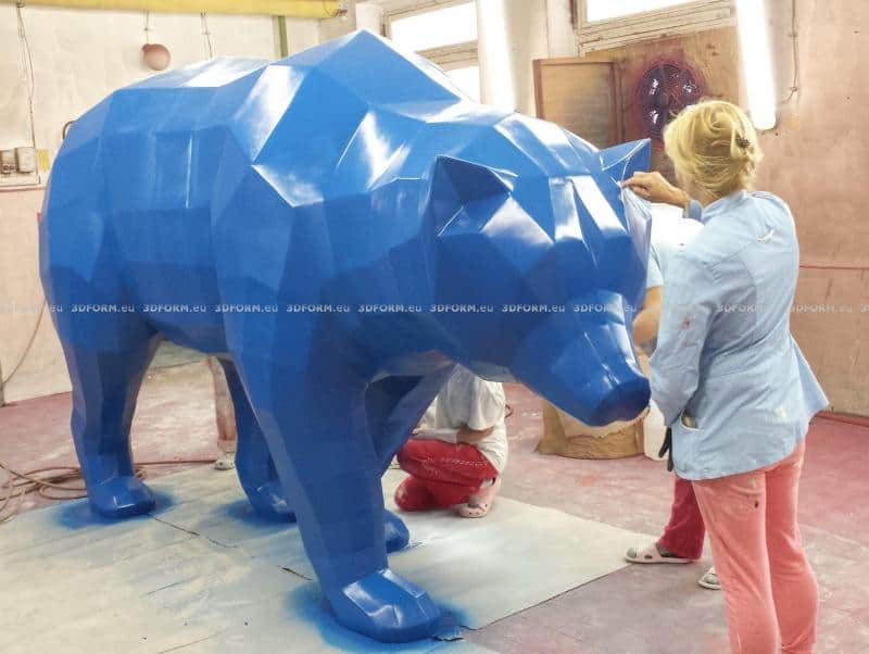 Geometrischer Low-Poly-Bär 1 Kobieta maluje dużą, niebieską, geometryczną rzeźbę niedźwiedzia w pracowni artystycznej.