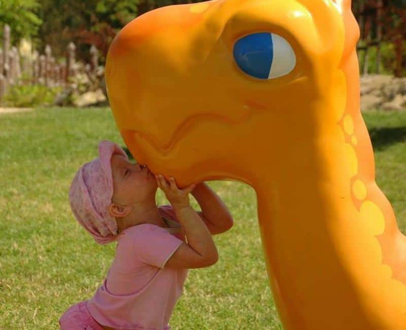 Drachen im Vergnügungspark 1 Dziewczynka w różowym ubraniu całuje dużą, pomarańczową figurkę dinozaura na placu zabaw.