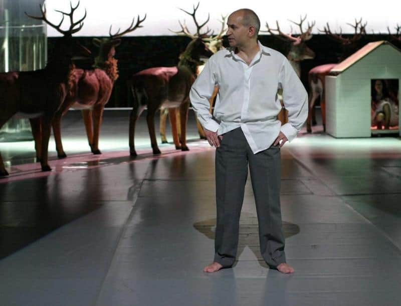 Bühnenbild-Elemente für das Dramatische Theater der Hauptstadt Warschau 1 Mężczyzna w białej koszuli i szarych spodniach stoi boso na scenie, w tle stoją wypchane jelenie.