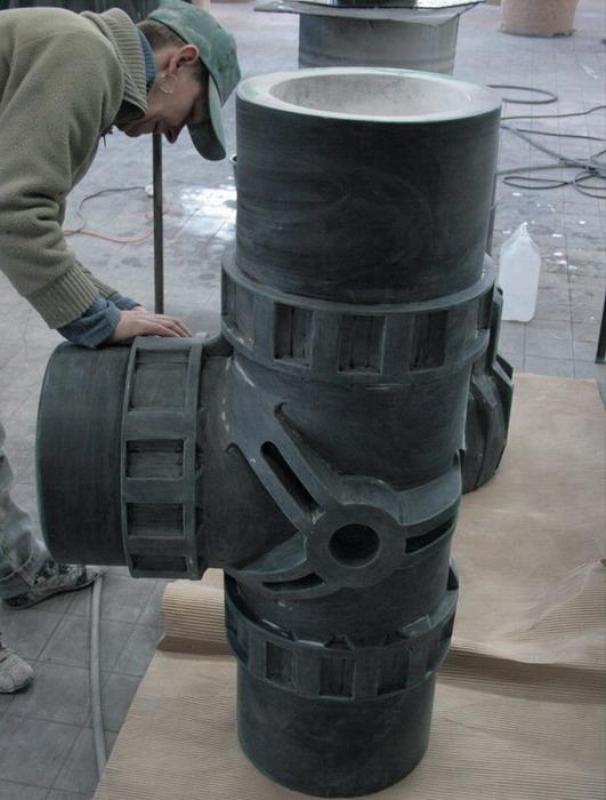 Mężczyzna w warsztacie pochylający się nad dużym rzeźbionym modelem krzyżowego połączenia rur.