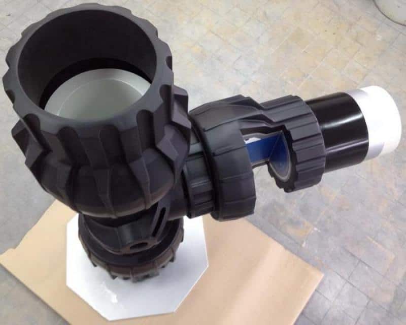 Czarny element mechaniczny o skomplikowanej budowie, z przegubem i uchwytem, na tle kartonowej powierzchni.