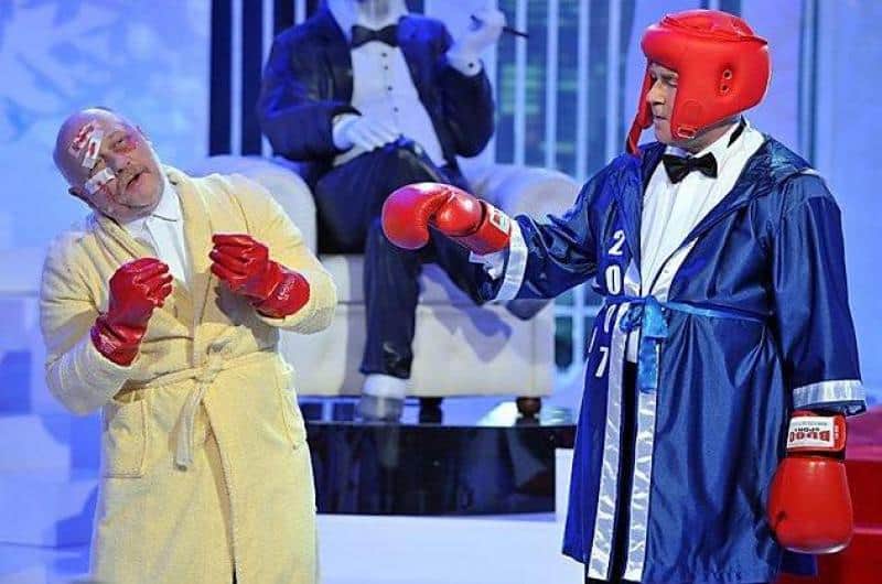 TVP2 - Bühnenelemente 2 Dwóch mężczyzn ubranych w stroje bokserskie i rękawice bokserskie podczas występu scenicznego, jeden w szlafroku, a drugi w niebieskim szlafroku bokserskim i kasku ochronnym.