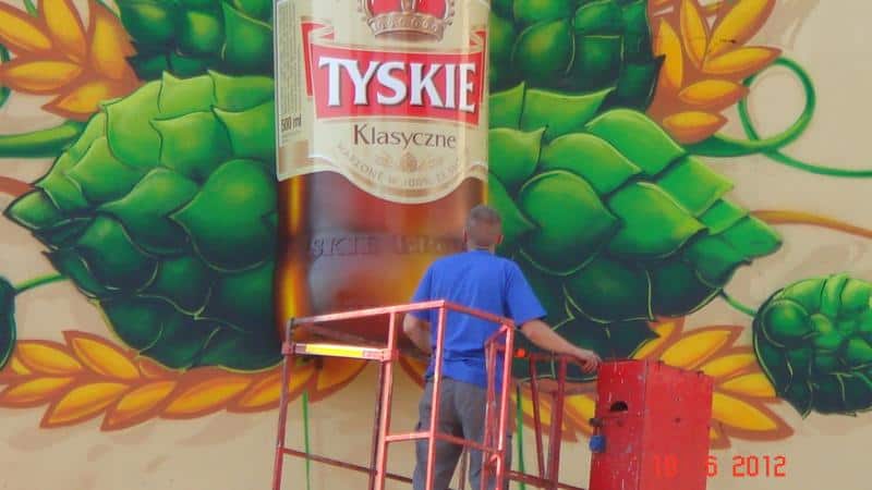 Dreidimensionales Wandbild TYSKIE 4 Mężczyzna na podnośniku malujący mural z butelką piwa Tyskie Klasyczne i chmielami.