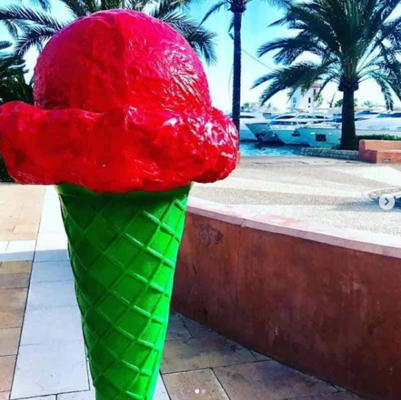 Wielka plastikowa rzeźba przedstawiająca loda rożka z czerwoną gałką na zielonym wafelku, umieszczona na zewnątrz w otoczeniu palm i jachtów.