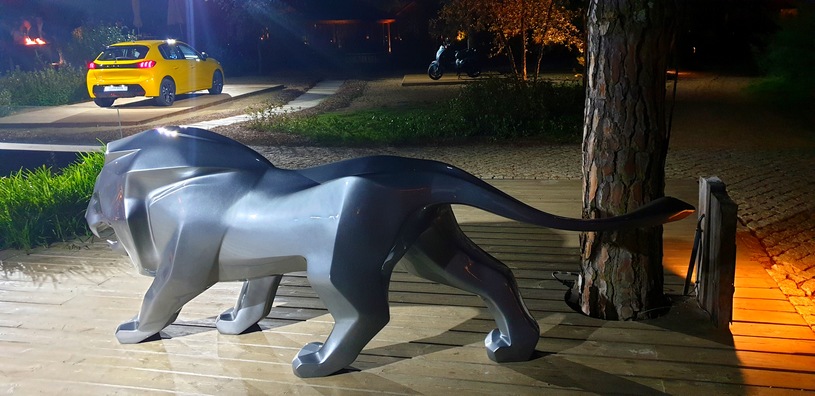 Löwe Peugeot 4 Figurka lwa w kolorze srebrnym na drewnianej platformie, w tle żółty samochód na oświetlonym podjeździe.