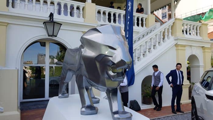 Löwe Peugeot 3 Metalowy posąg lwa na tle białego budynku z kolumnadą i schodami, przy którym stoją elegancko ubrani ludzie.