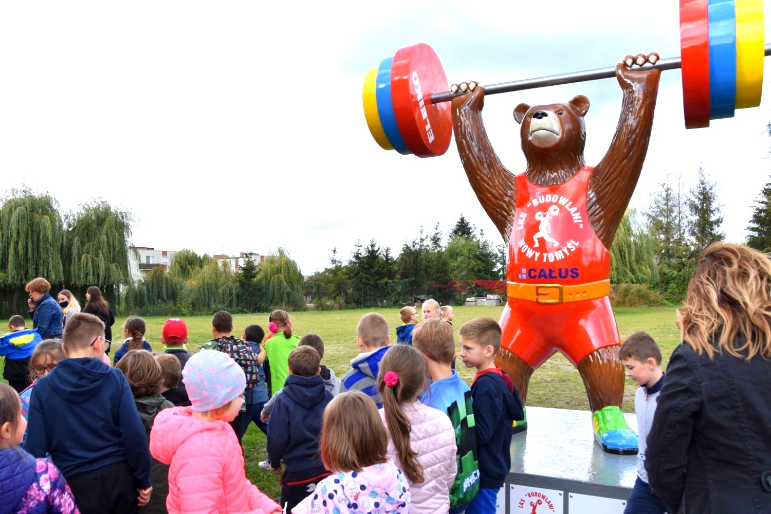 Großer Bär - Gewichtheben-Meisterschaften 4 Dzieci z nauczycielkami oglądają dużą figurę misia podnoszącego sztangę na zewnątrz.