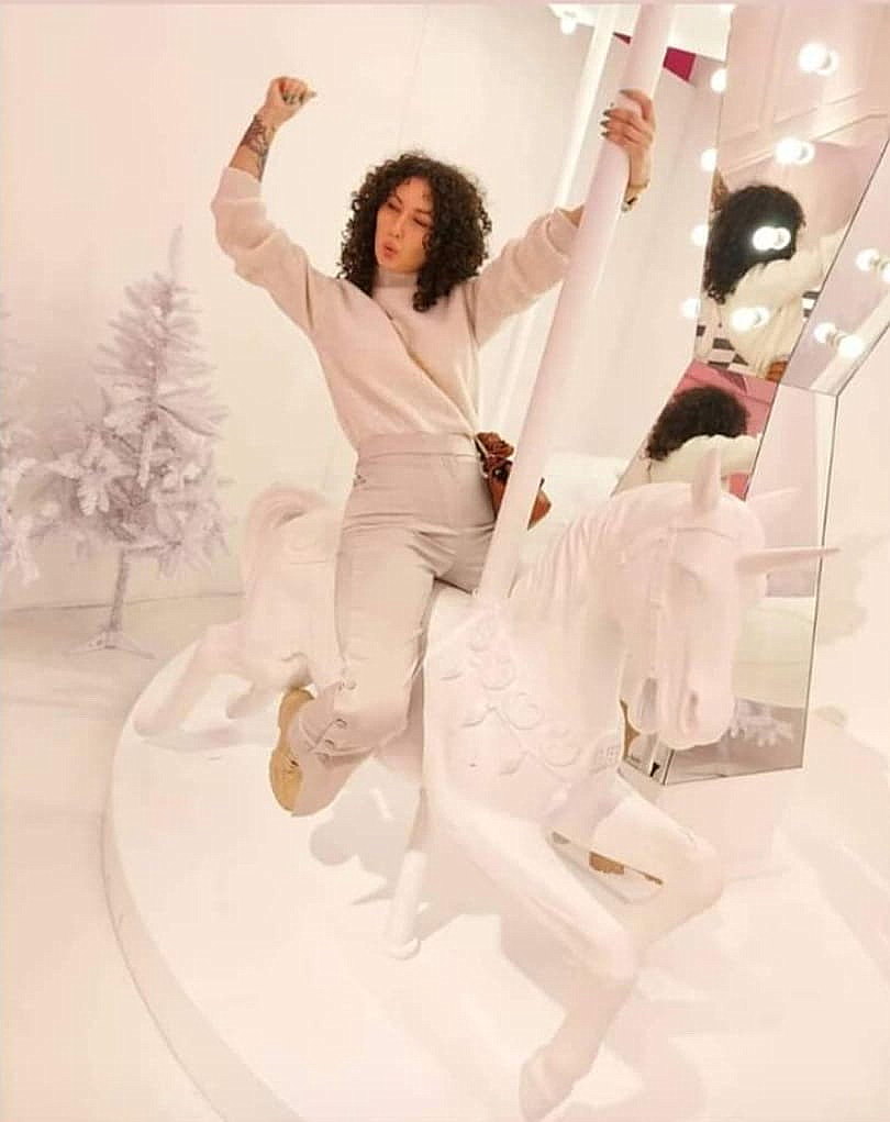 Osoba siedząca na karuzeli z białym jednorożcem w jasnym pomieszczeniu, obok znajdują się lustra i ozdobione białe choinki.