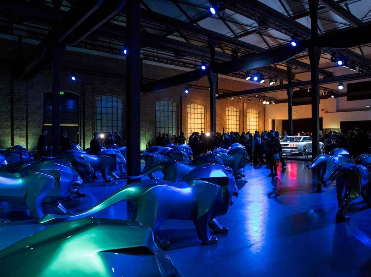 Löwe Peugeot 2 Pomieszczenie wypełnione niebieskimi rzeźbami lwów, z ludźmi i samochodem w tle, oświetlone niebieskim i fioletowym światłem.