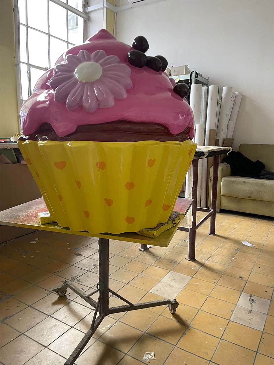 Bunter Cupcake für die Konditorei Sweet Home in Lagos, Portugal. 2 Duża, kolorowa rzeźba muffinki w żółtej papilotce z serduszkami, z różowym lukrem, ozdobiona kwiatkiem i wiśniami, umieszczona na stole w pracowni.