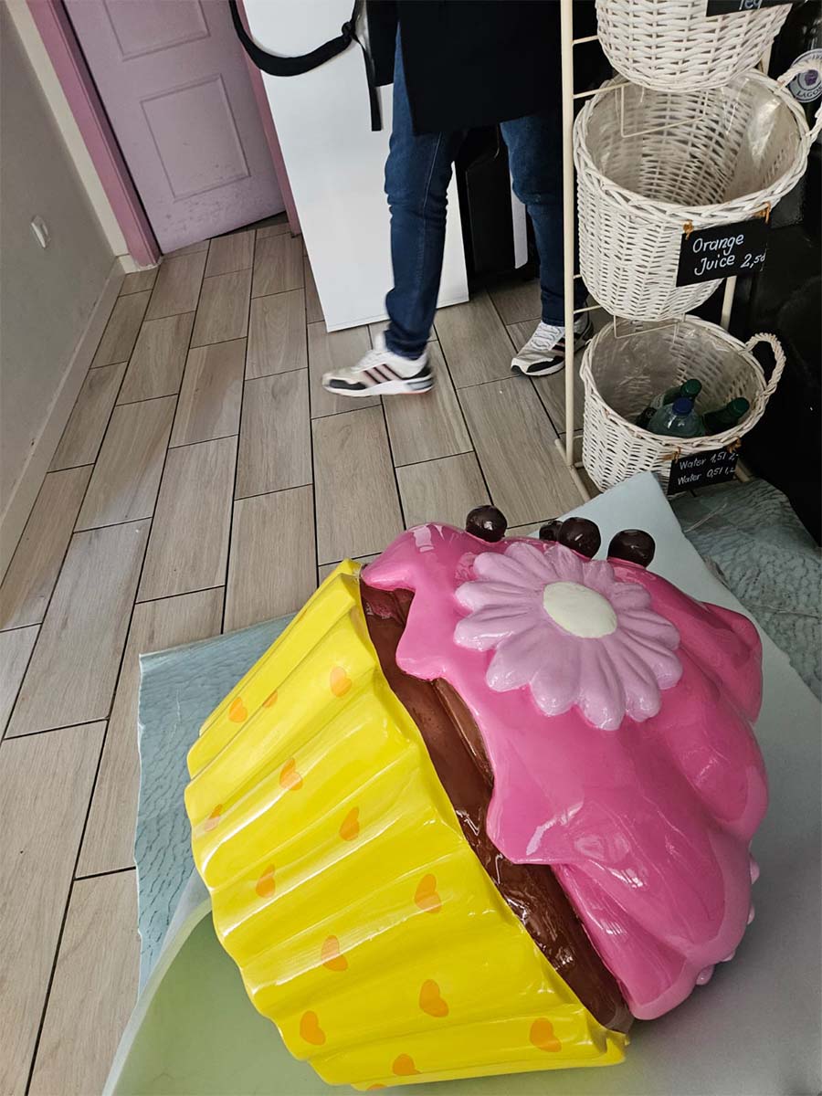 Bunter Cupcake für die Konditorei Sweet Home in Lagos, Portugal. 3 Rzeźba wielkiego babeczki w pastelowych kolorach stojąca na podłodze w kawiarni obok osoby w dżinsach i białych butach.