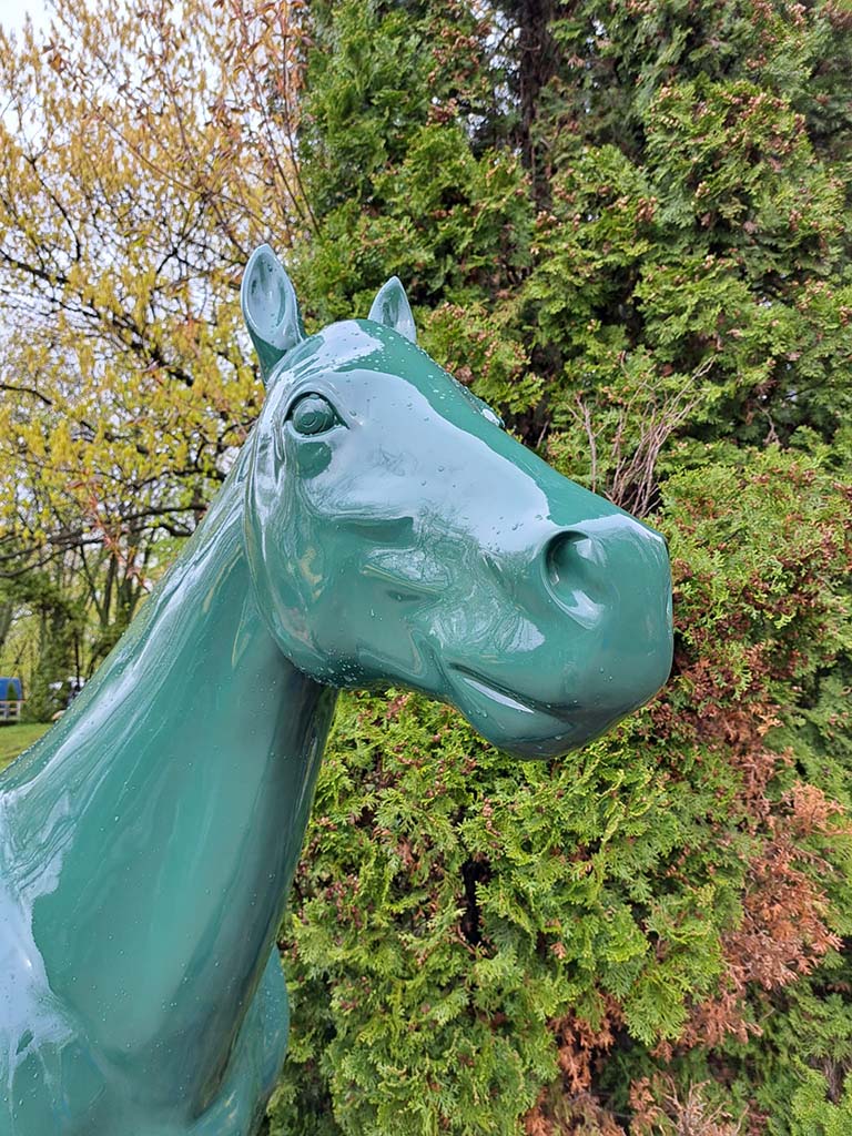 Bunte Pferdeskulpturen auf dem Parcours Partynice in Warschau 6 Zielona rzeźba konia na tle drzew.