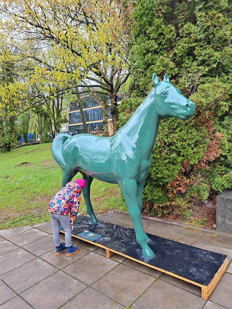 Bunte Pferdeskulpturen auf dem Parcours Partynice in Warschau 4 Dziecko w kolorowej kurtce zaglądające pod rzeźbę zielonego konia na zewnątrz w parku.