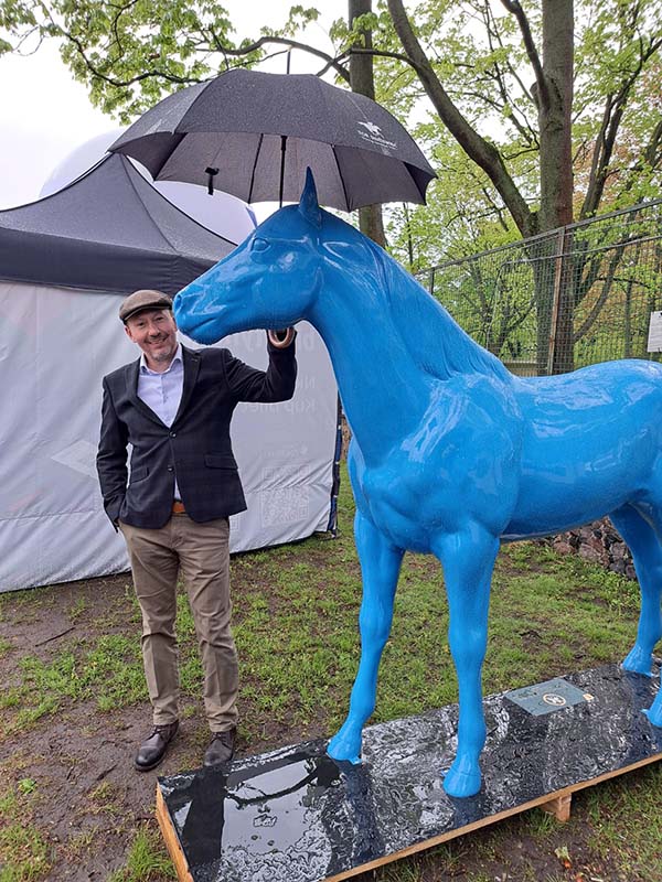 Bunte Pferdeskulpturen auf dem Parcours Partynice in Warschau 2 Mężczyzna trzymający parasol nad niebieską statuetką konia.