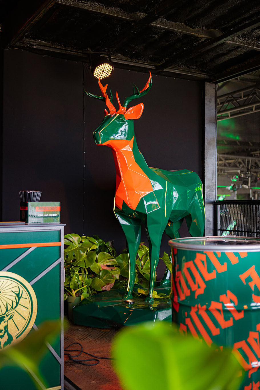 Pomarańczowo-zielona rzeźba jelenia stojąca w otoczeniu roślin i zielono-pomarańczowych elementów dekoracyjnych.