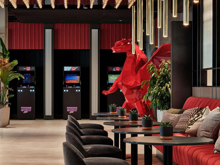 Wnętrze nowoczesnego salonu z czerwoną ławą, czarnymi fotelami, stylowymi stołami i dużą czerwoną rzeźbą smoka, w tle automaty do gier retro.