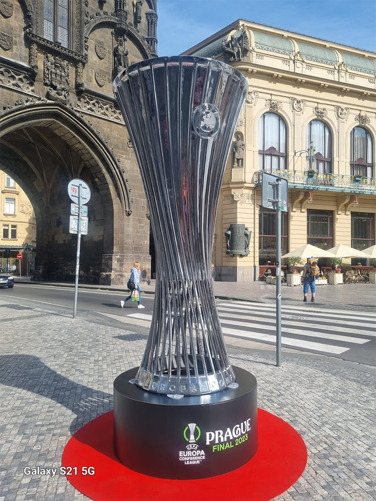 Replika pucharu UEFA Europa Conference League na tle historycznych budynków w Pradze z napisem PRAGUE FINAL 2023 na podstawie pucharu ustawionego na czerwonym dywanie.