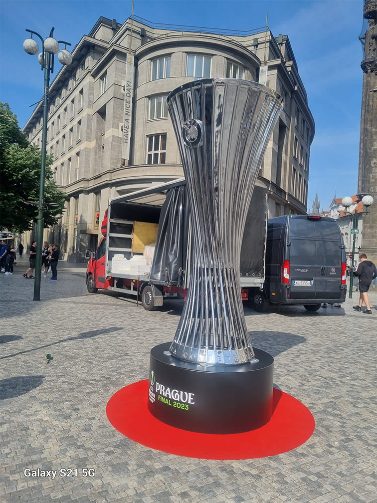 Duża replika trofeum UEFA Europa Conference League na cokole z napisem PRAGUE FINAL 2023 ustawiona na czerwonym okręgu na brukowanym placu w otoczeniu budynków i samochodów dostawczych w słoneczny dzień