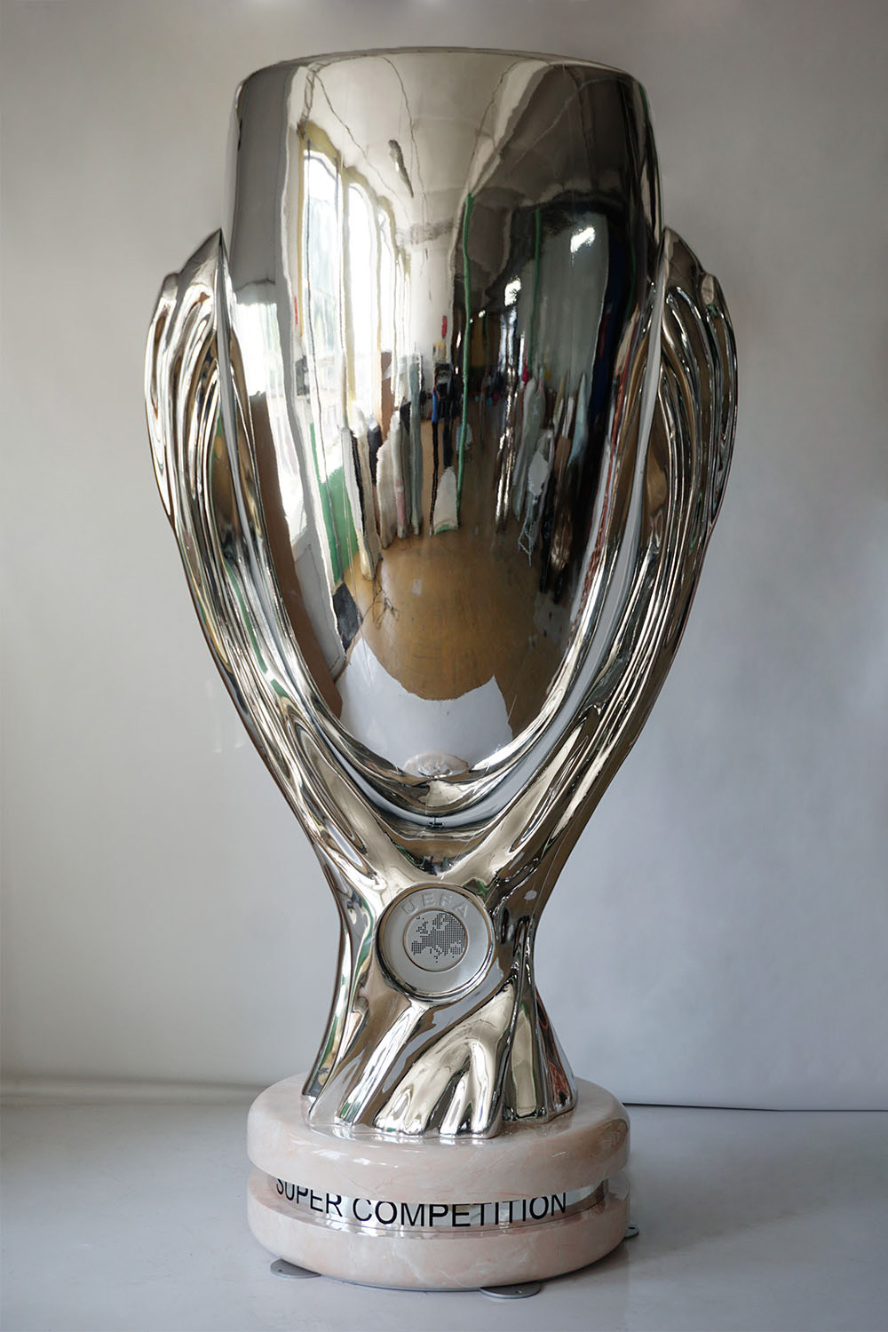 Połyskujący srebrny puchar UEFA Super Competition stojący na jasnym marmurowym podstawkach na białym tle.