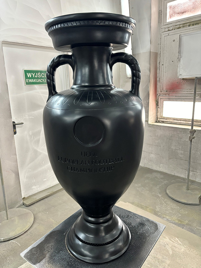 Duży czarny model trofeum UEFA European Football Championship ustawiony w pomieszczeniu z białymi ścianami i dużym oknem.