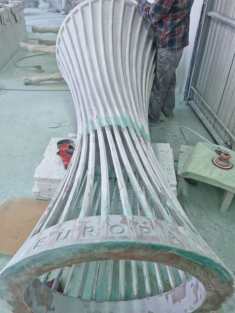 Dużych rozmiarów rzeźba w trakcie obróbki, wykonana z materiału kamieniopodobnego, o smukłym, zakrzywionym kształcie z napisem EUROPA na dolnej części, w warsztacie ze szlifierkami i narzędziami wokół, częściowo zamazany biały i zielonkawy kolor.