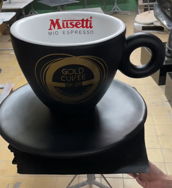 Werbefiguren der Musetti-Tassen 1 Duża czarna filiżanka kawy z napisem Musetti Mio Espresso na białym wnętrzu oraz złotym napisem Gold Cuvée 1994-2014 na zewnątrz, ustawiona na czarnym spodku, na tle pomieszczenia z jasną podłogą i stołami.