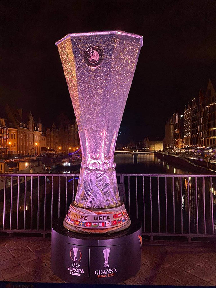 Replika trofeum UEFA Europa League na czarnym postumencie z napisem Gdańsk Final 2021, ustawiona na tle nocnej panoramy miasta z oświetlonymi zabytkowymi budynkami i wodą w tle
