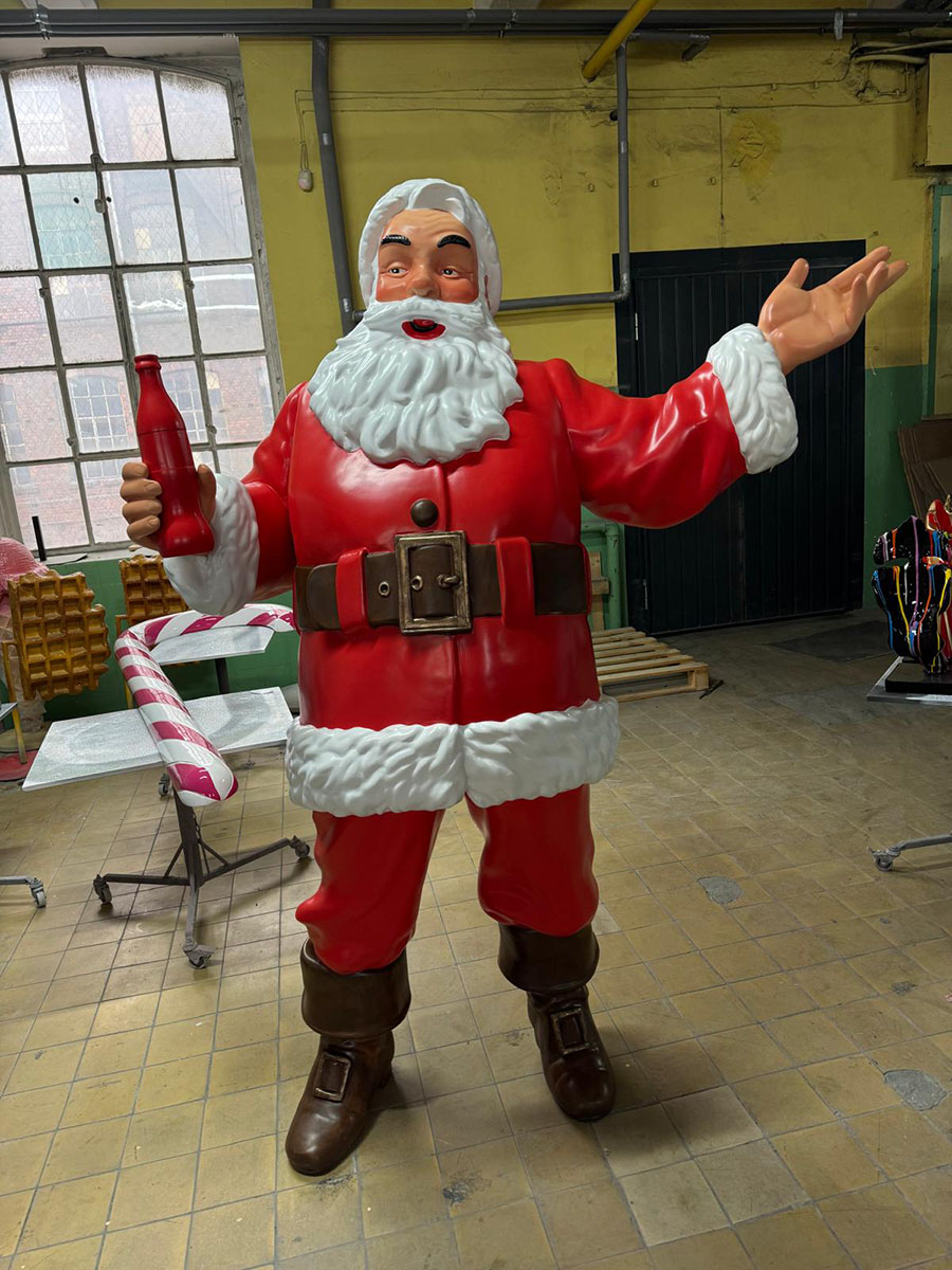 Jeder kann der Weihnachtsmann sein: Coca-Colas Weihnachtskampagne mit Weihnachtsmannfiguren 17 Figurka Świętego Mikołaja w czerwonym stroju i brązowych butach stojąca na podłodze w pomieszczeniu z żółto-zielonymi ścianami i dużym oknem, trzymająca czerwoną butelkę w jednej ręce i gestykulująca drugą.