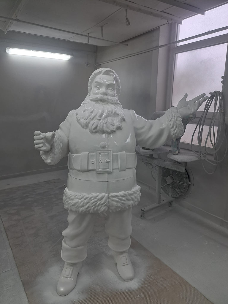 Jeder kann der Weihnachtsmann sein: Coca-Colas Weihnachtskampagne mit Weihnachtsmannfiguren 18 Biały, niepomalowany posąg Świętego Mikołaja w pracowni malarskiej, z uniesionymi rękoma, obok okno i wentylator.