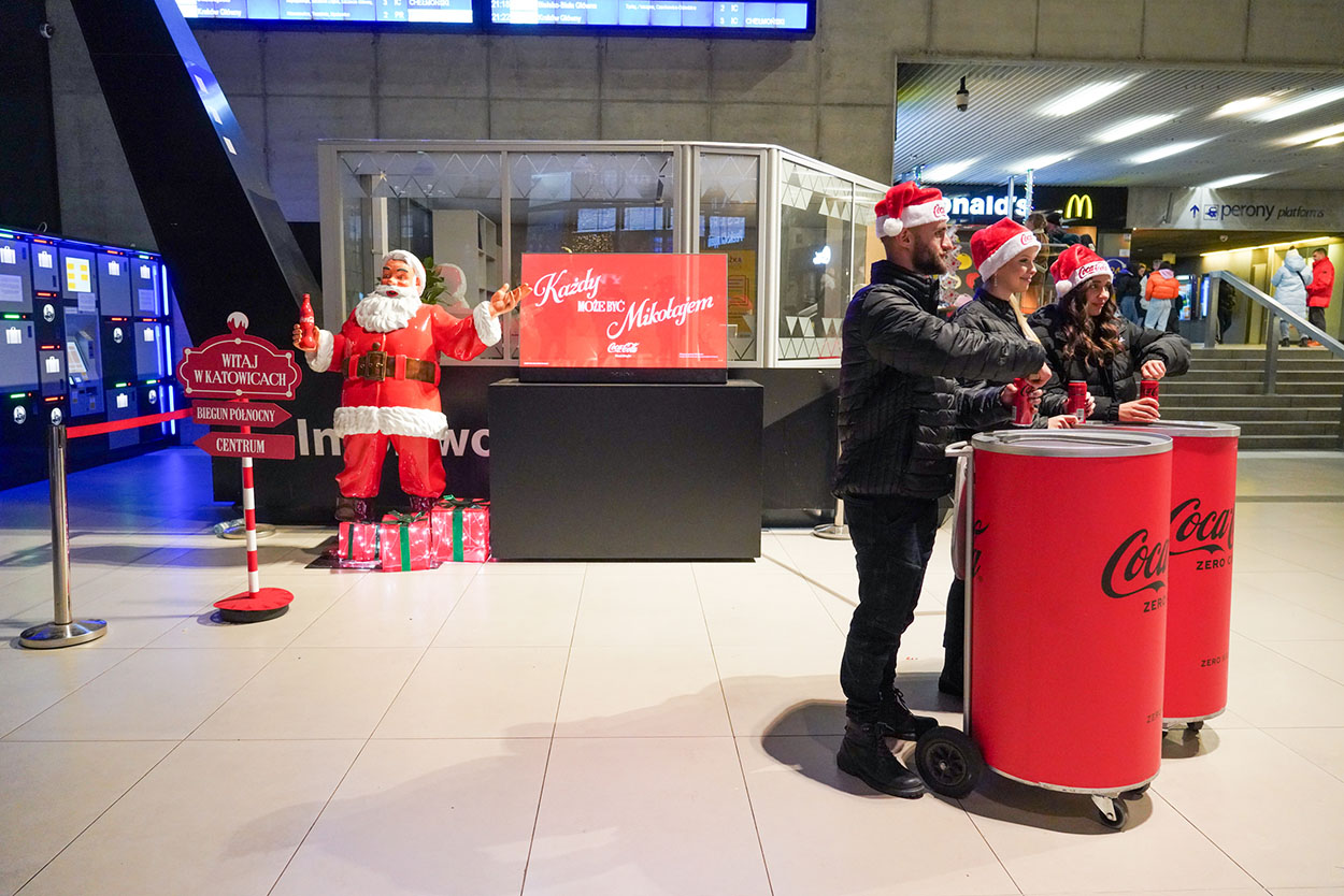 Jeder kann der Weihnachtsmann sein: Coca-Colas Weihnachtskampagne mit Weihnachtsmannfiguren 3 Trzy osoby w czapkach św. Mikołaja stoją przy czerwonych pojemnikach Coca-Coli w hali dworca, obok stoi figura św. Mikołaja z butelką Coca-Coli i znakiem