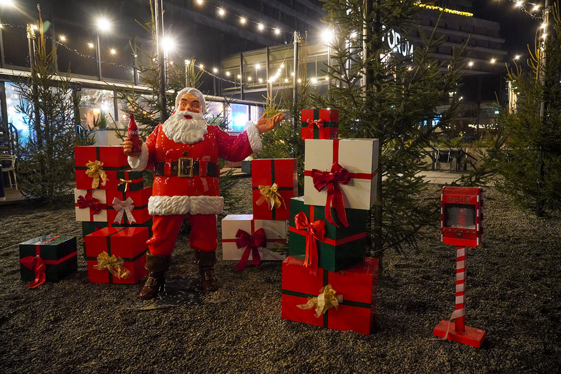 Jeder kann der Weihnachtsmann sein: Coca-Colas Weihnachtskampagne mit Weihnachtsmannfiguren 1 Figurka Świętego Mikołaja trzymającego butelkę Coca-Coli, otoczona licznymi prezentami w czerwonych, białych i zielonych opakowaniach z kokardami, ustawiona na żwirowym podłożu na tle drzewek iglastych i oświetlenia świątecznego nocą